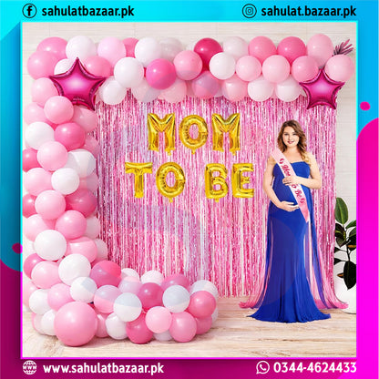 Woman in a blue dress with a 'Mom to Be' sash standing in front of a pink and white balloon arch with 'Mom to Be' balloons.